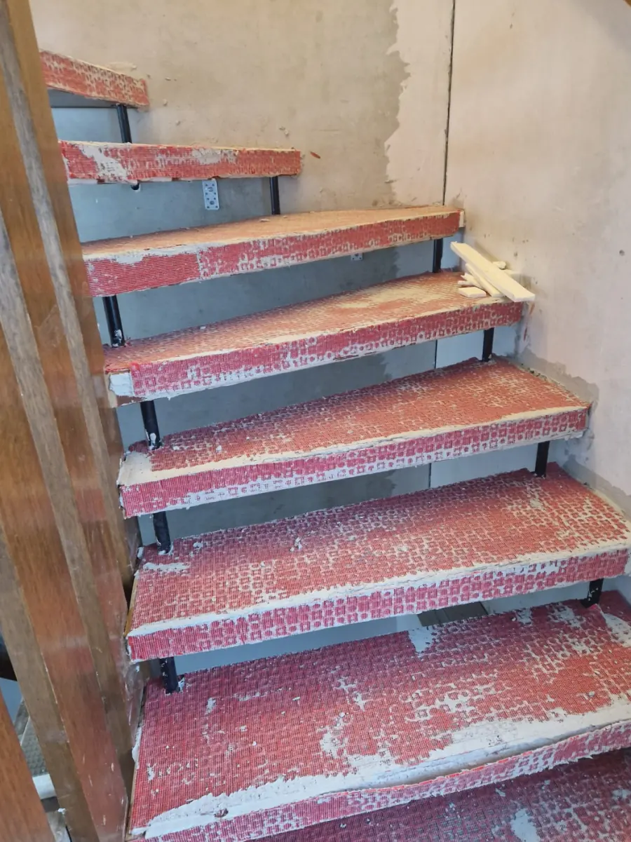 Treppe vor dem Umbau: rohe Stufen mit Resten von altem Belag und Kleber, schwarze Stahlhalter darunter, links das Holzgeländer